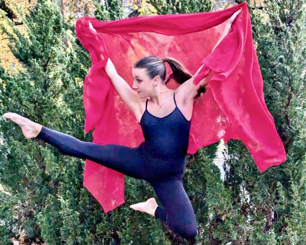 Olga Dunn dancer leaping with red cape outside in front of greenery